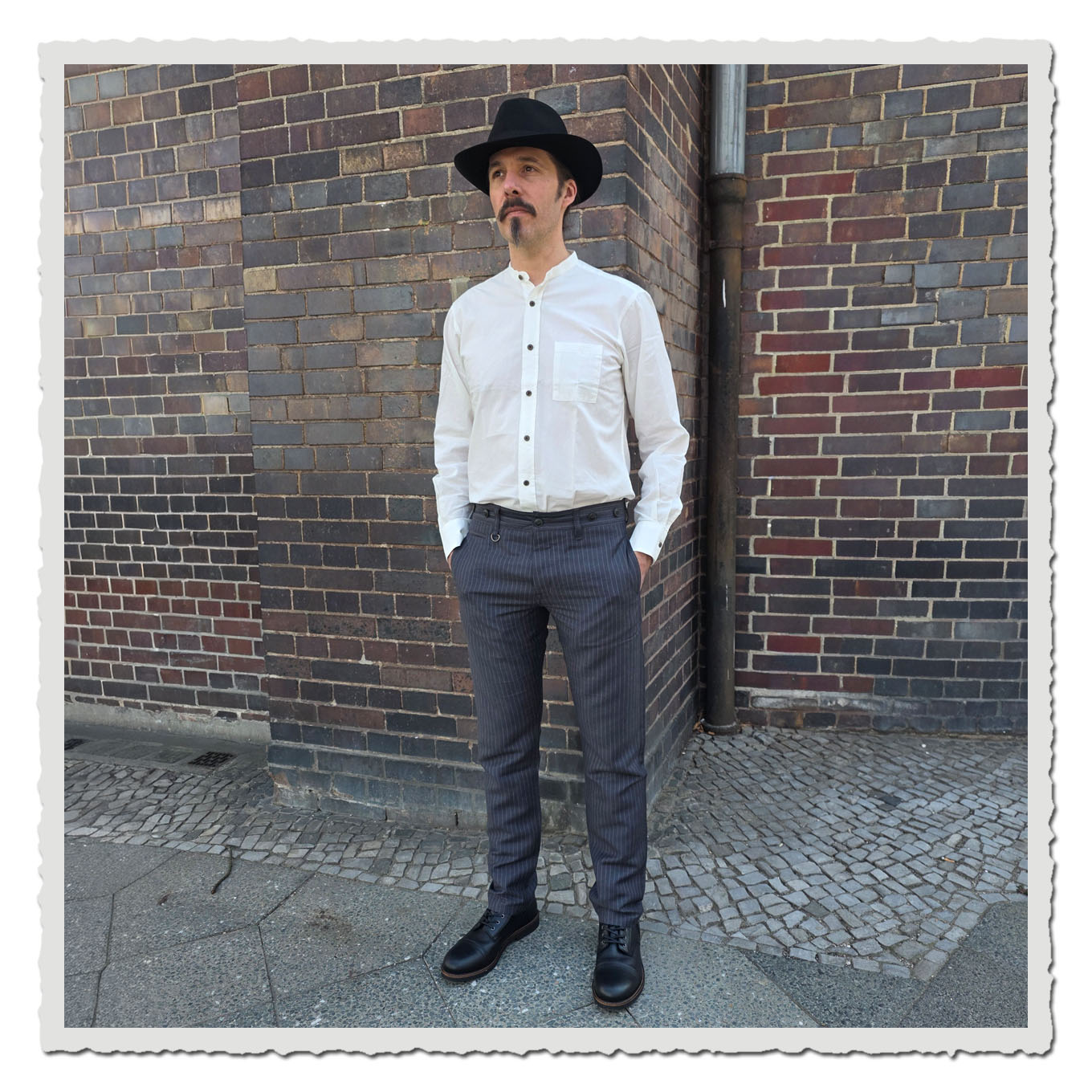 Ganzkörperansicht eines Models mit grauer Nadelstreifenhose vor dunkler Backsteinwand im Außenbereich.